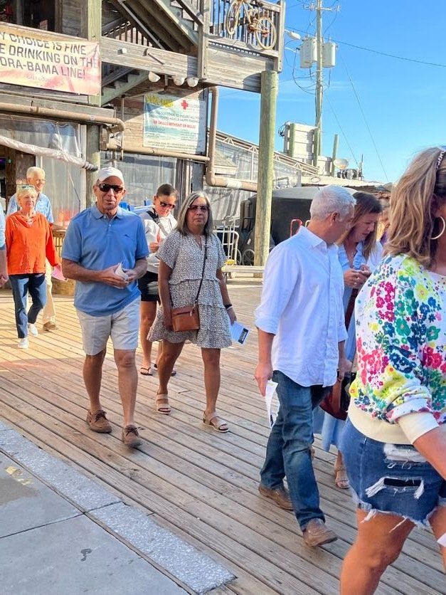 People walk on a wooden boardwalk near the Flora-Bama lounge on a sunny day.
