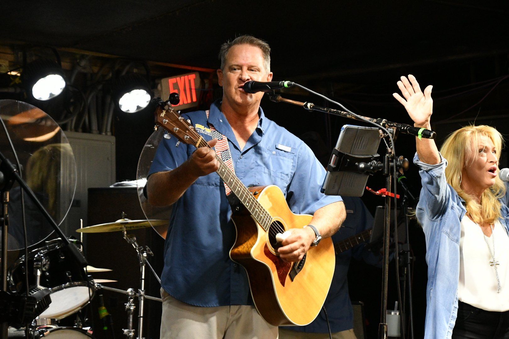 A man playing an acoustic guitar and singing into a microphone next to a person with one arm raised on a dimly lit stage.