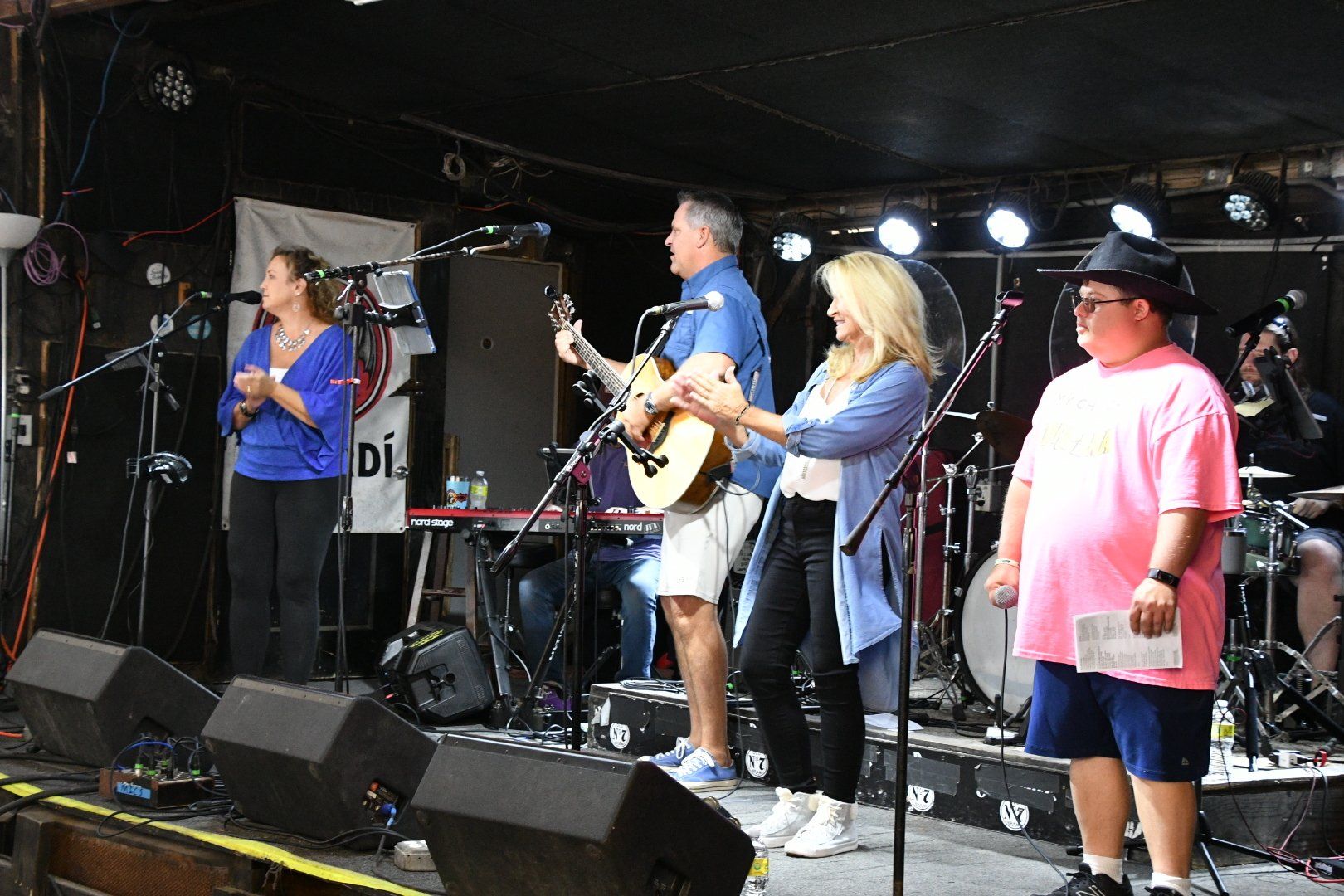 A band performs on stage with a singer, guitarist, vocalist, and a person in a pink shirt and black hat holding a paper.