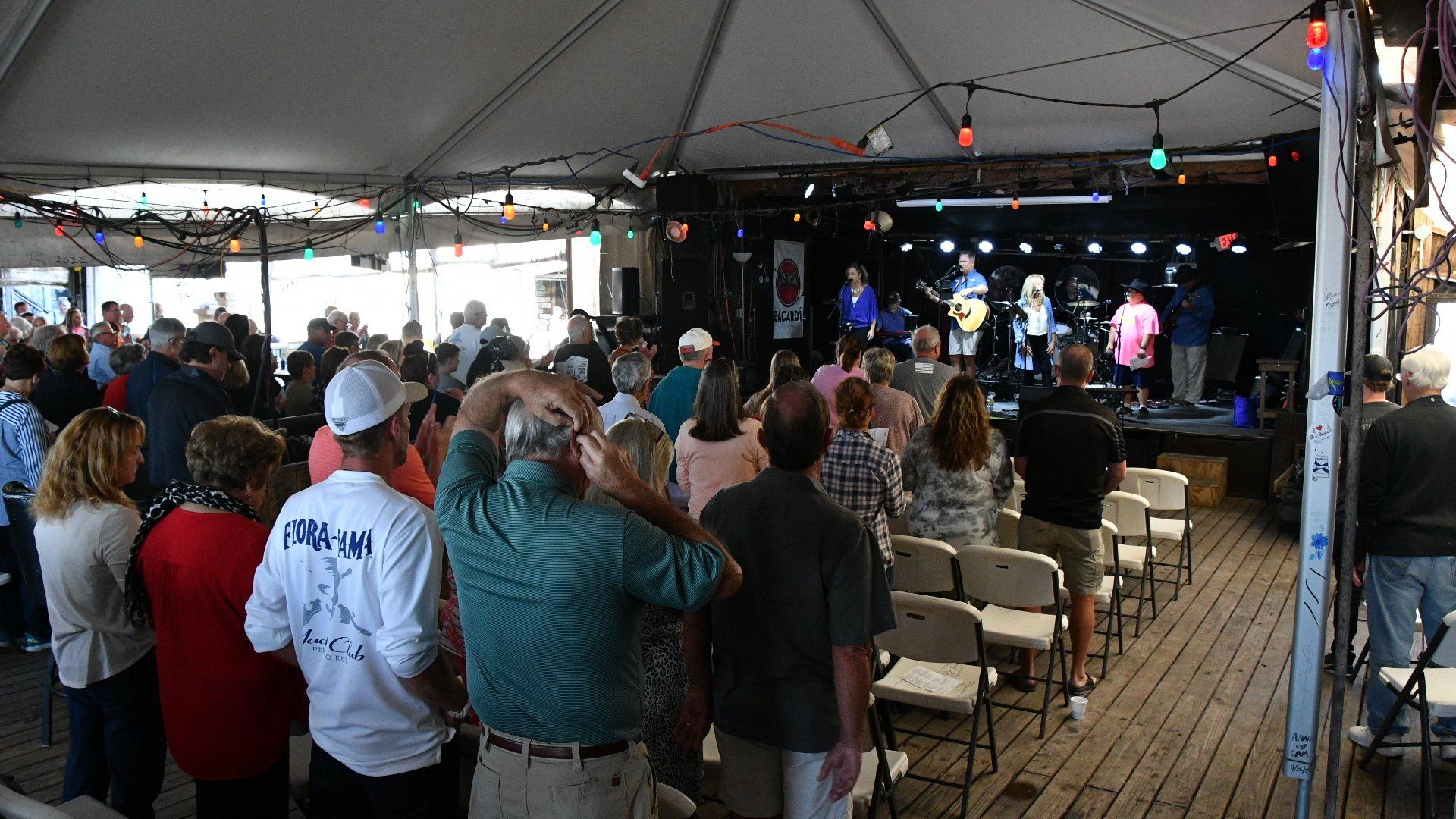 A crowd watches a musical performance under a tent, with people standing and sitting in chairs facing a lit stage.