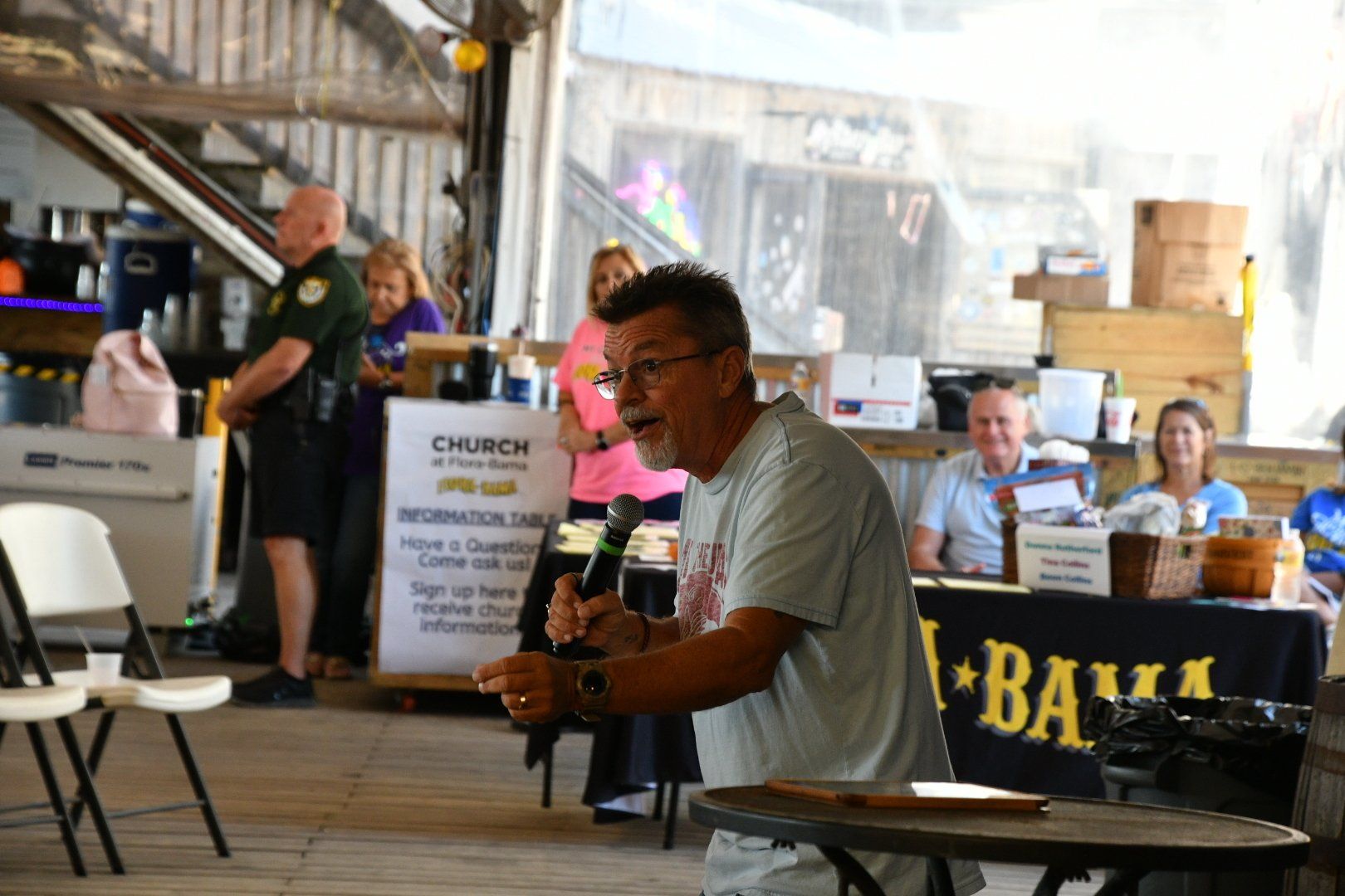 A man holding a microphone speaks to an audience in a covered outdoor pavilion with a 