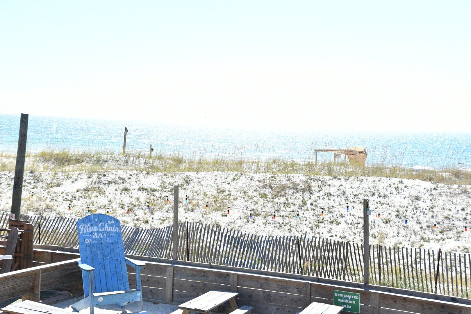 A blue chair sits on a wooden deck in front of a sandy beach, dunes, and the ocean under a bright sky.