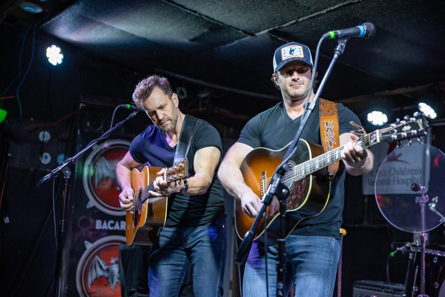 Two musicians play acoustic guitars on stage, both wearing black shirts and blue jeans, performing under stage lighting.