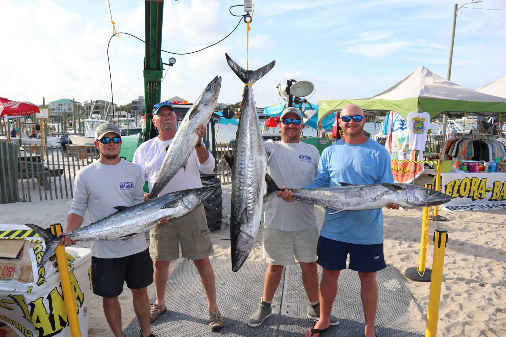 Four people stand on a sandy beach holding four large fish caught during a fishing competition at a dockside venue.
