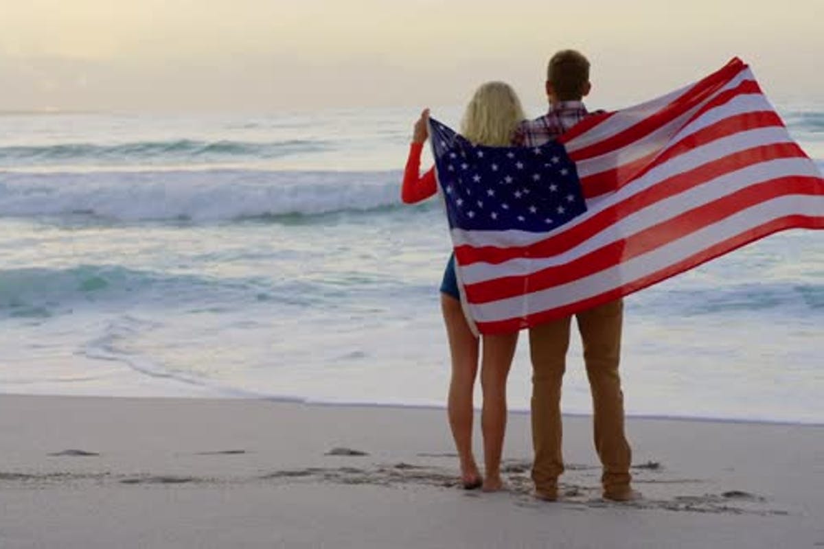 Two people stand on a beach, facing the ocean while wrapped together in an American flag at sunset.