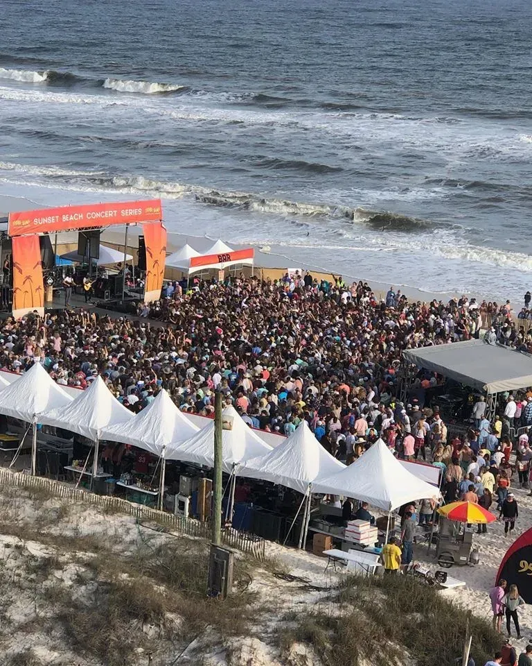 A crowded beach concert with white tents lined up, facing a large stage near the ocean shoreline on a sunny day.