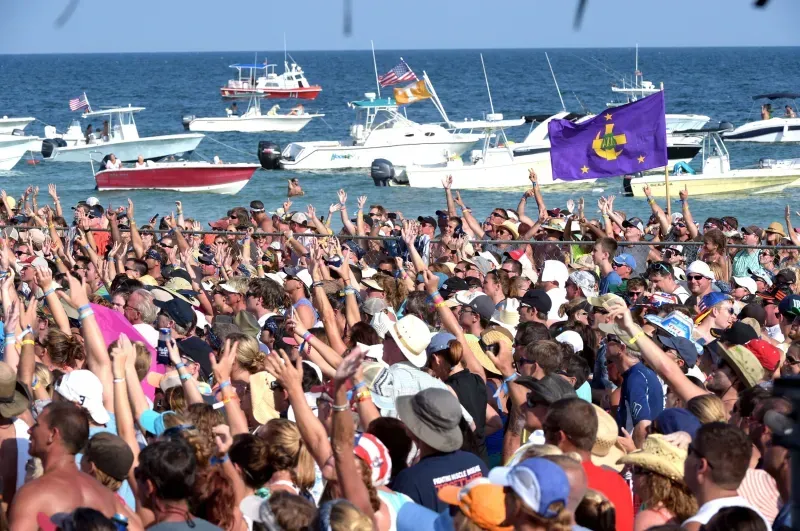 A large, cheering crowd stands on a beach facing the ocean, filled with many boats, under a bright, sunny sky.