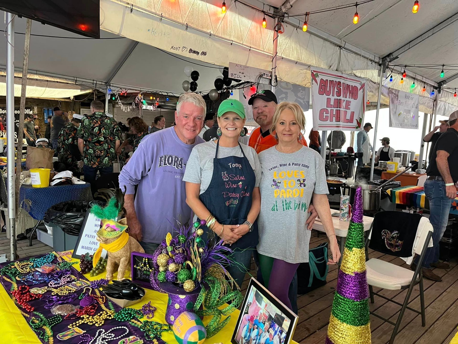 Three people pose behind a table decorated with Mardi Gras beads and colors under a tent at an outdoor festival.