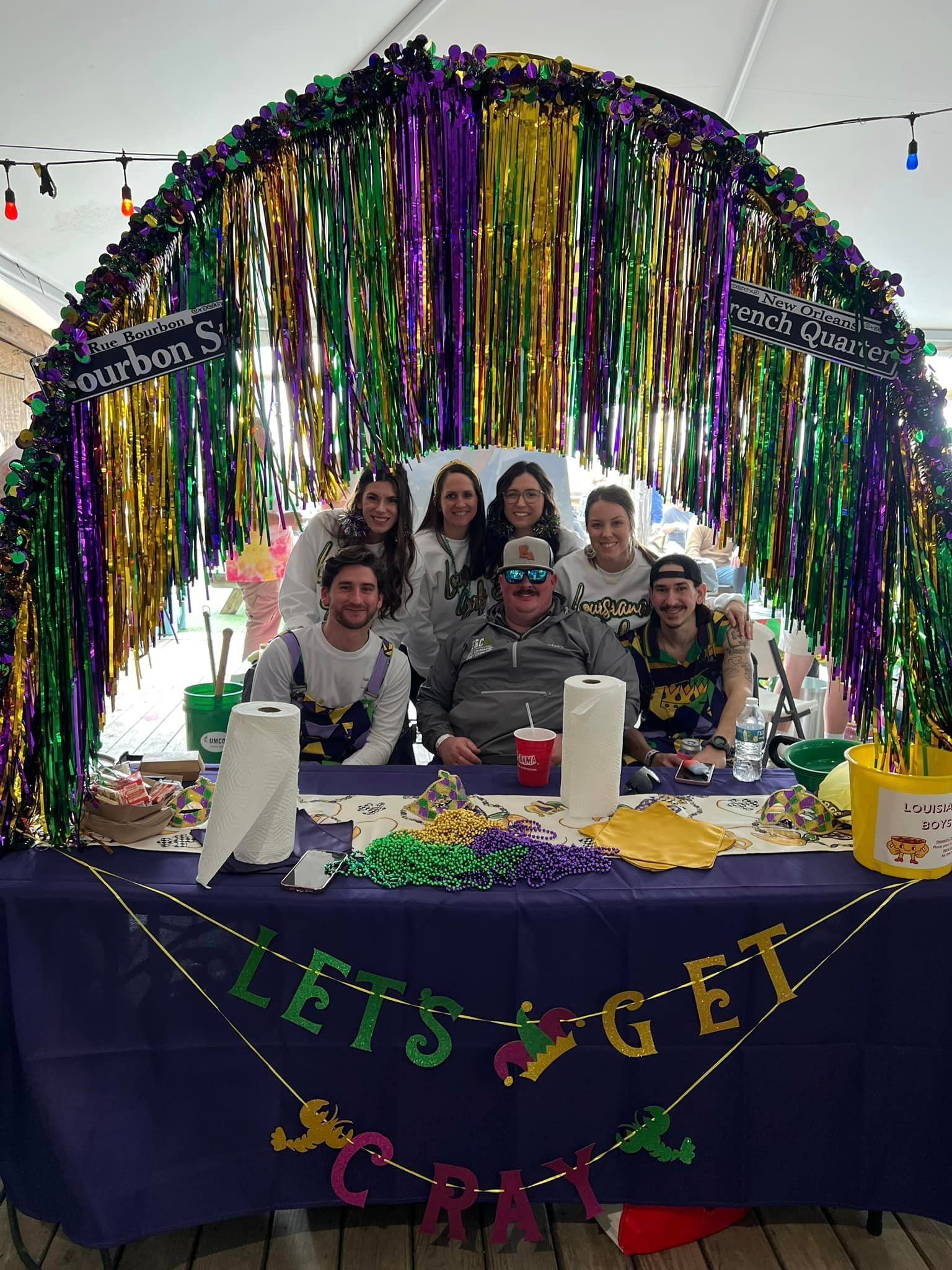 Seven people posing behind a table decorated with Mardi Gras colors, a tinsel archway, and a 