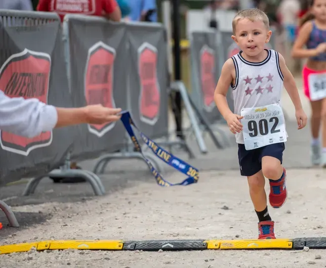 A child in a white tank top with stars running toward a finish line, with a race medal being held out to them.