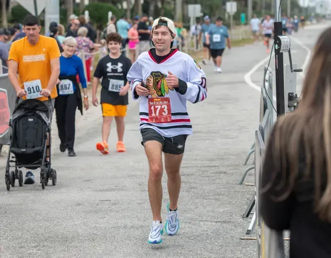 A runner wearing a white Chicago Blackhawks hockey jersey and number 173 bib runs toward the camera on a paved path.