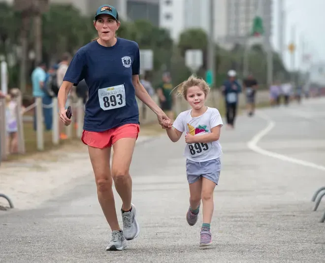 An adult and a child running side-by-side on a road during an outdoor race, both wearing race bibs and holding hands.