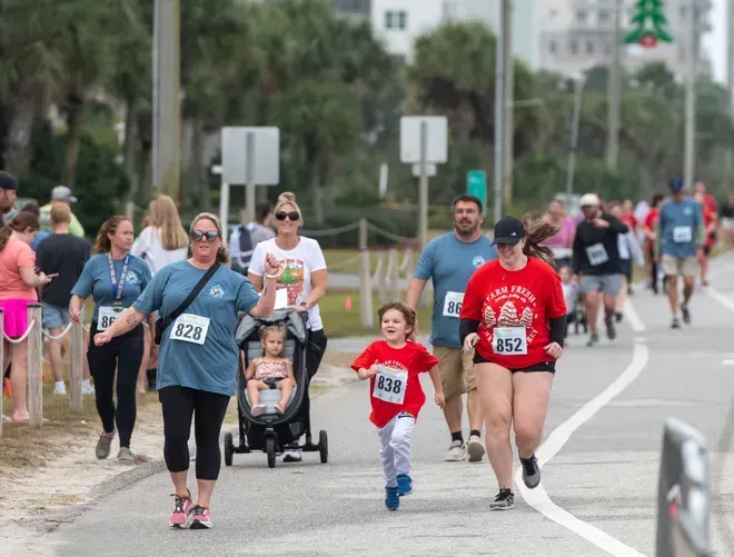 A group of people running on a paved path during an outdoor race, with some participants wearing numbered race bibs.