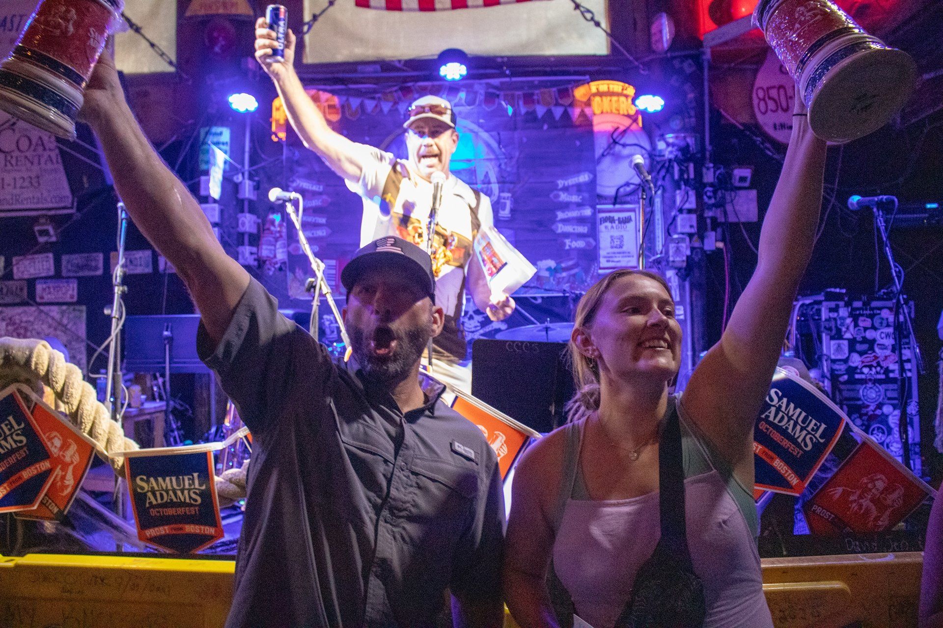 A man and woman raise beer steins in a dimly lit bar, with a musician performing on a stage in the background.