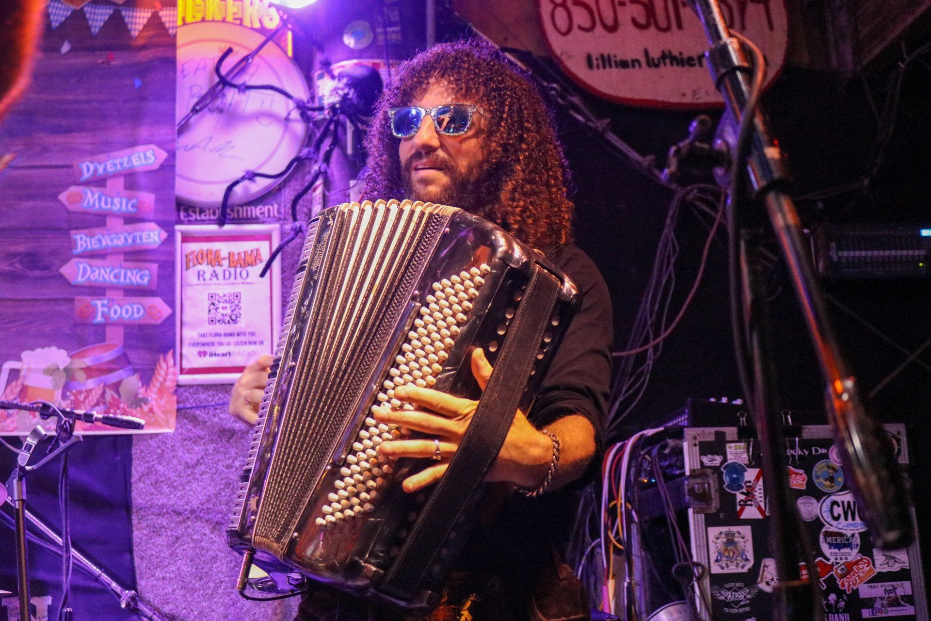 A person with curly hair and sunglasses plays a piano accordion on a dimly lit stage with purple lighting.