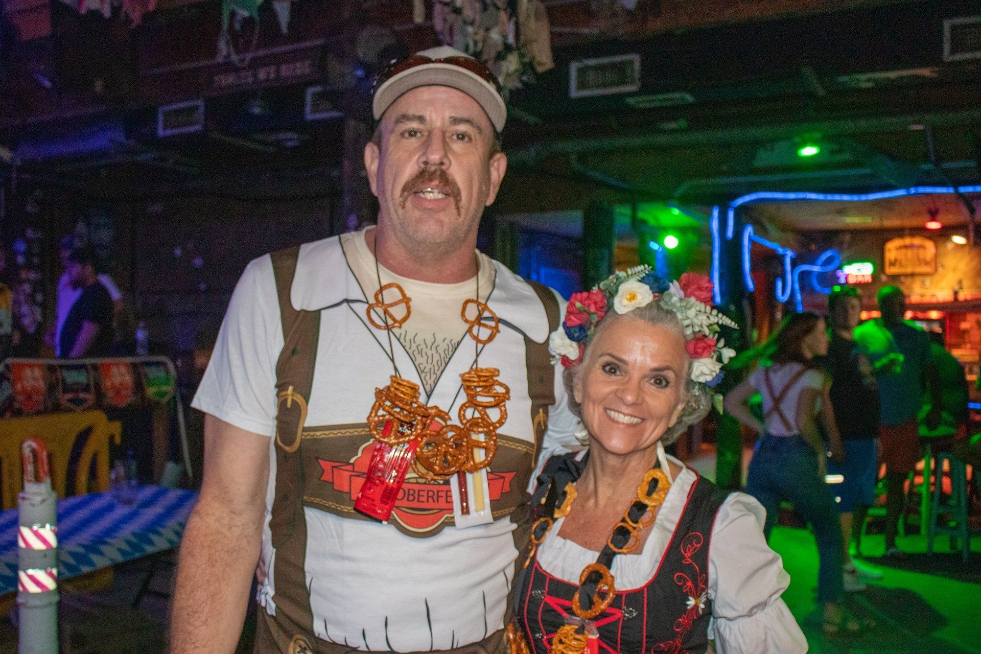 A person with a pretzel necklace and a person in a floral crown smiling in a rustic bar setting.