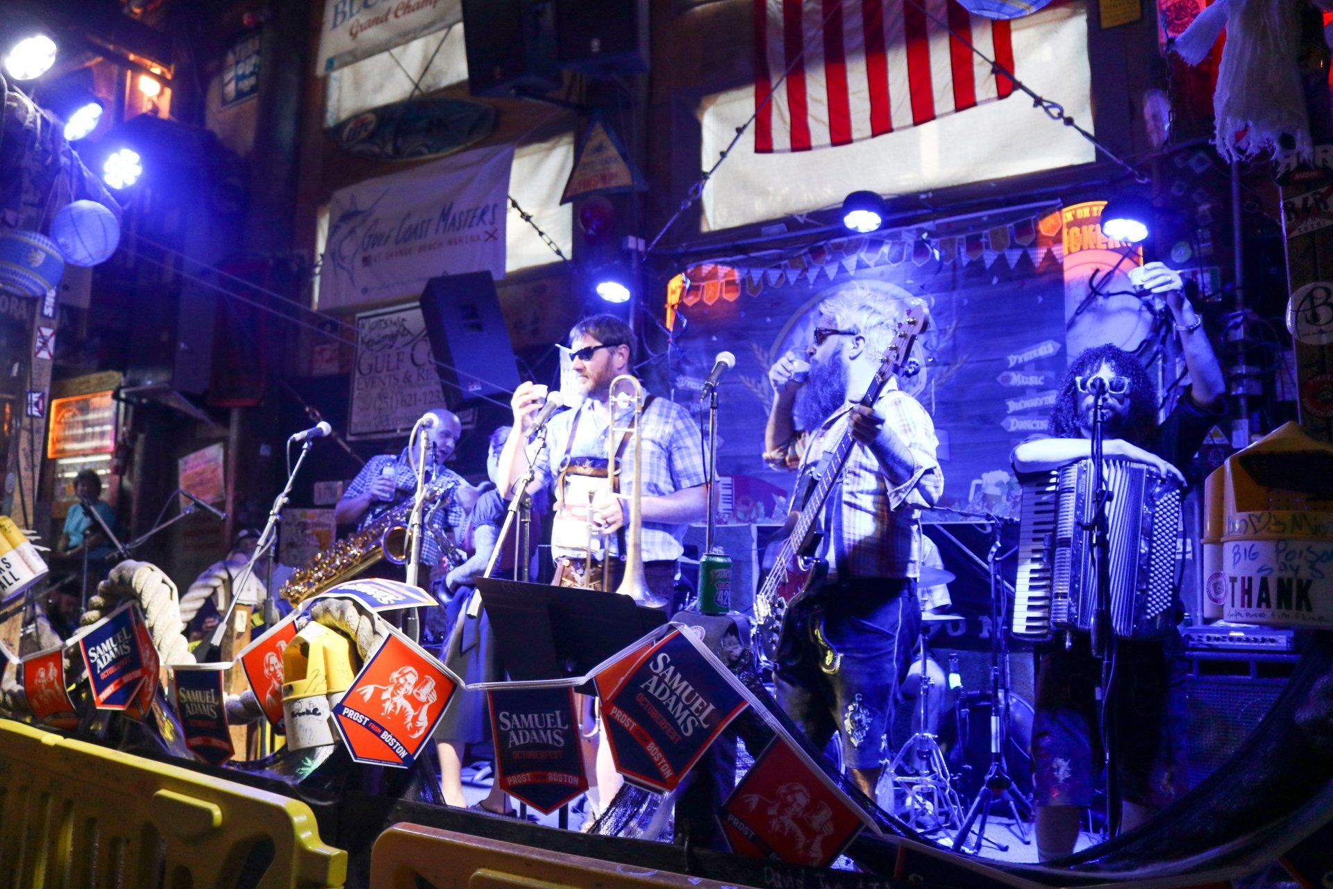 A musical band performs on a dimly lit stage under blue lighting, decorated with flags and an American flag in the back.