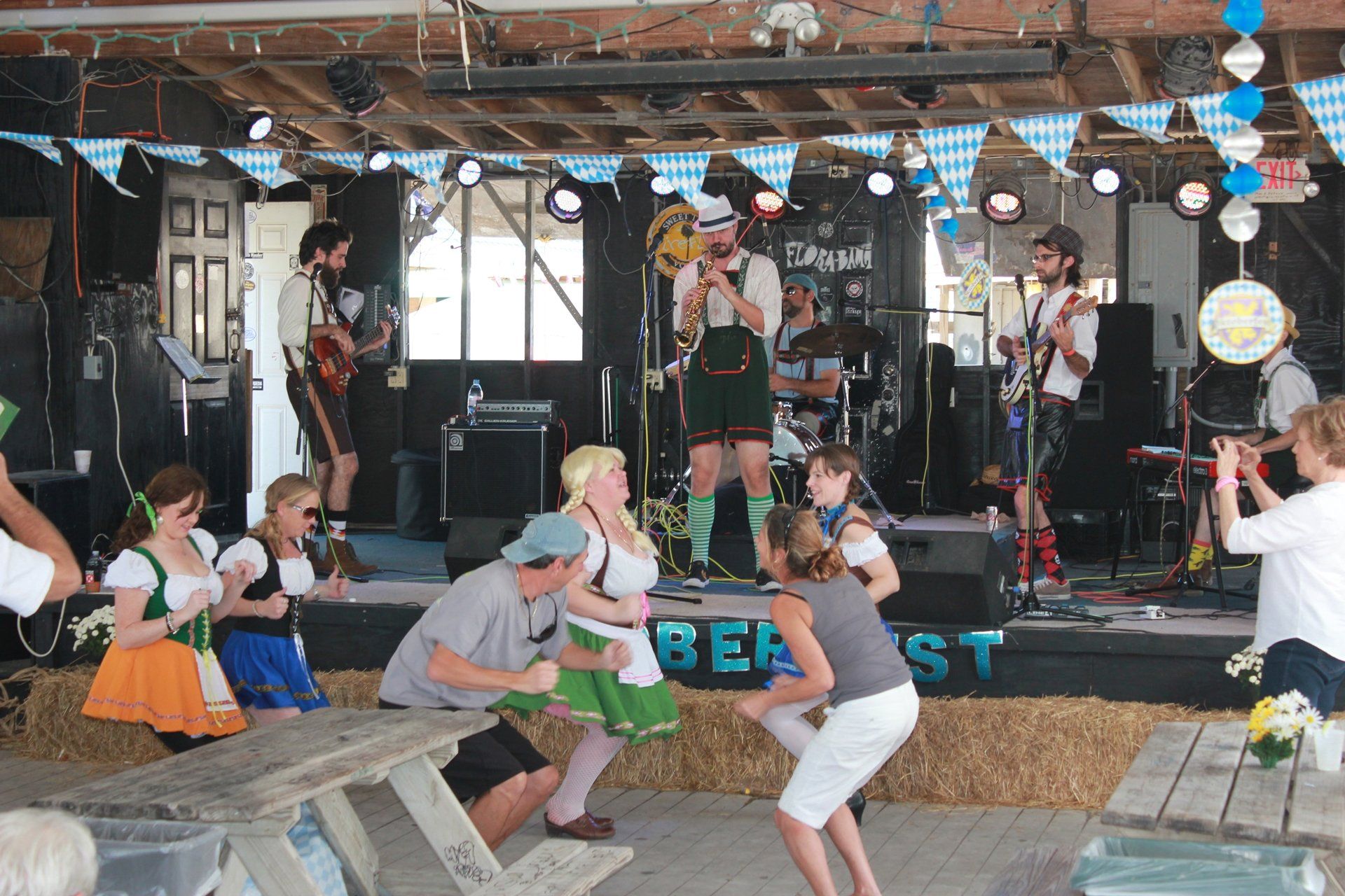 A band performs on a stage decorated with Oktoberfest signage, as people dance in front in traditional German attire.