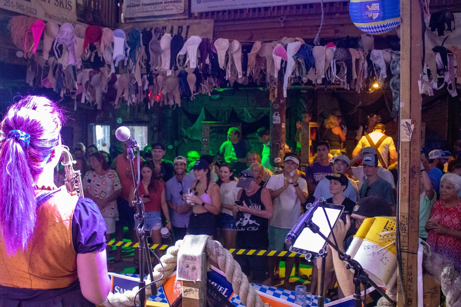 A crowd watches a performer on stage in a rustic venue, with dozens of bras hanging from the ceiling rafters.