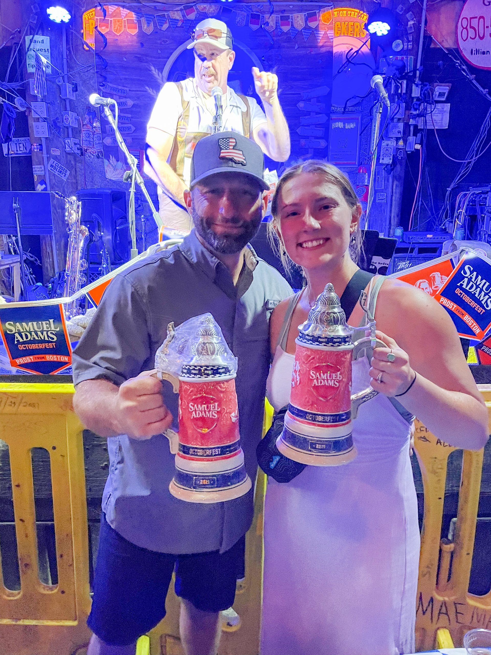 A smiling man and woman pose in a music venue, holding matching beer steins in front of a performing musician.