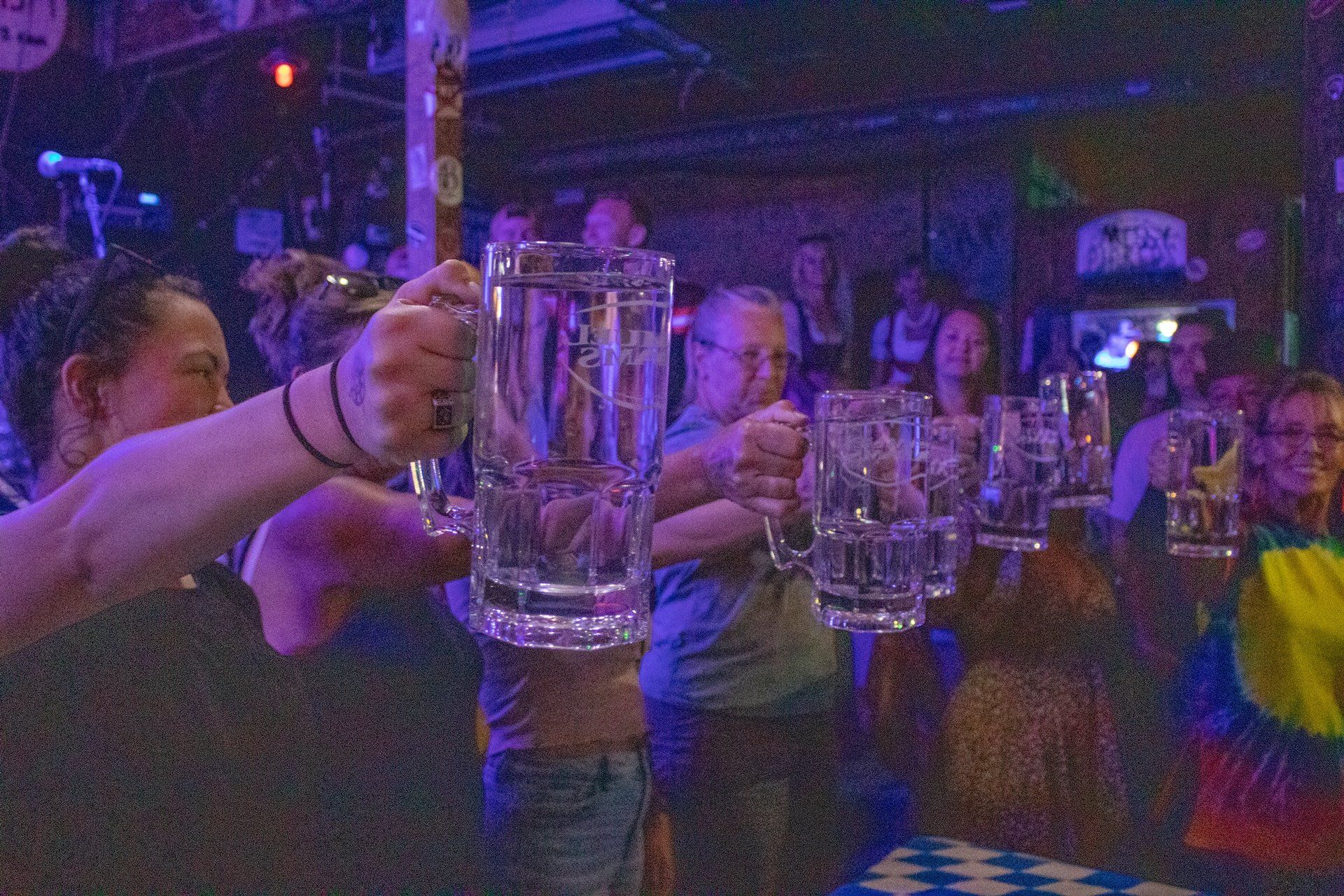 People in a dimly lit bar hold up large glass beer mugs in a toast.