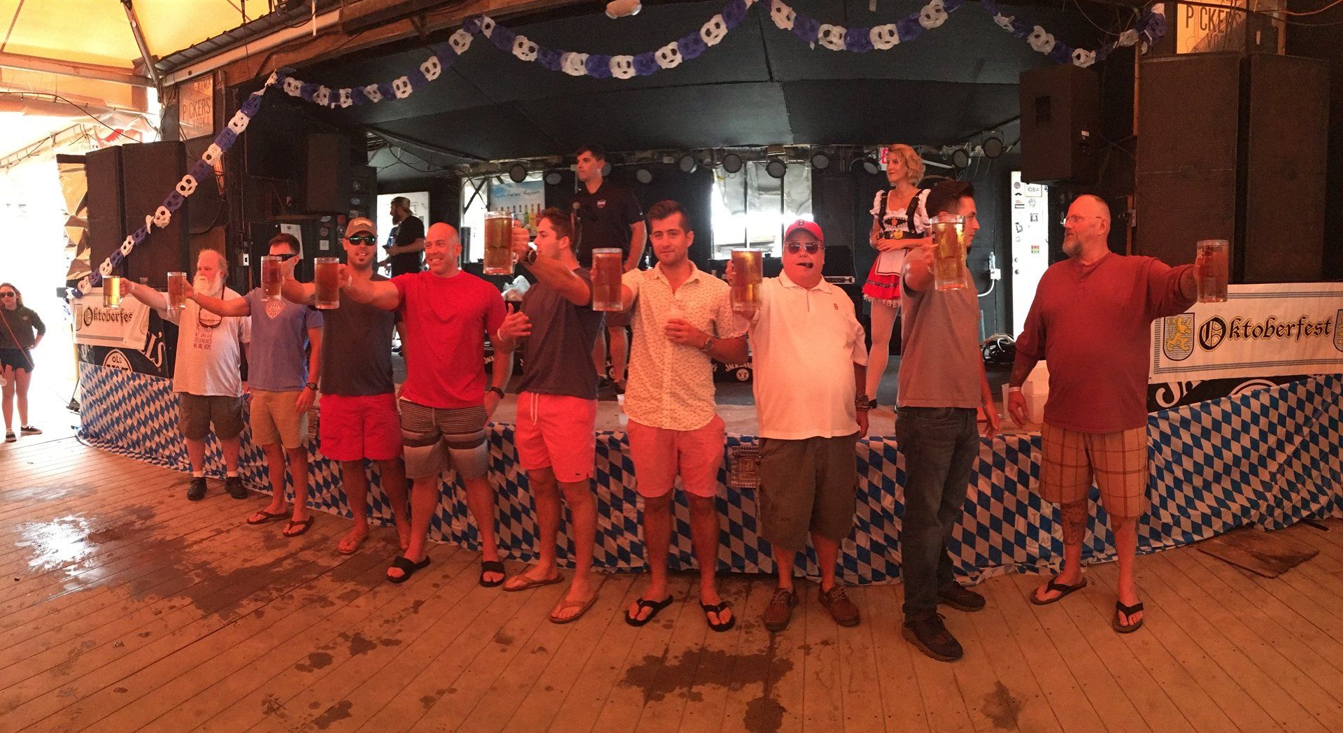 A group of people standing on a stage at an Oktoberfest-style event, holding up beer steins in a toast.