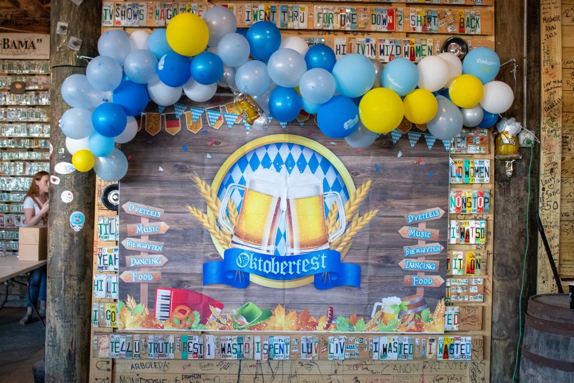Oktoberfest backdrop with a beer mug graphic and balloon arch, set against a wood-paneled wall decorated with license plates.