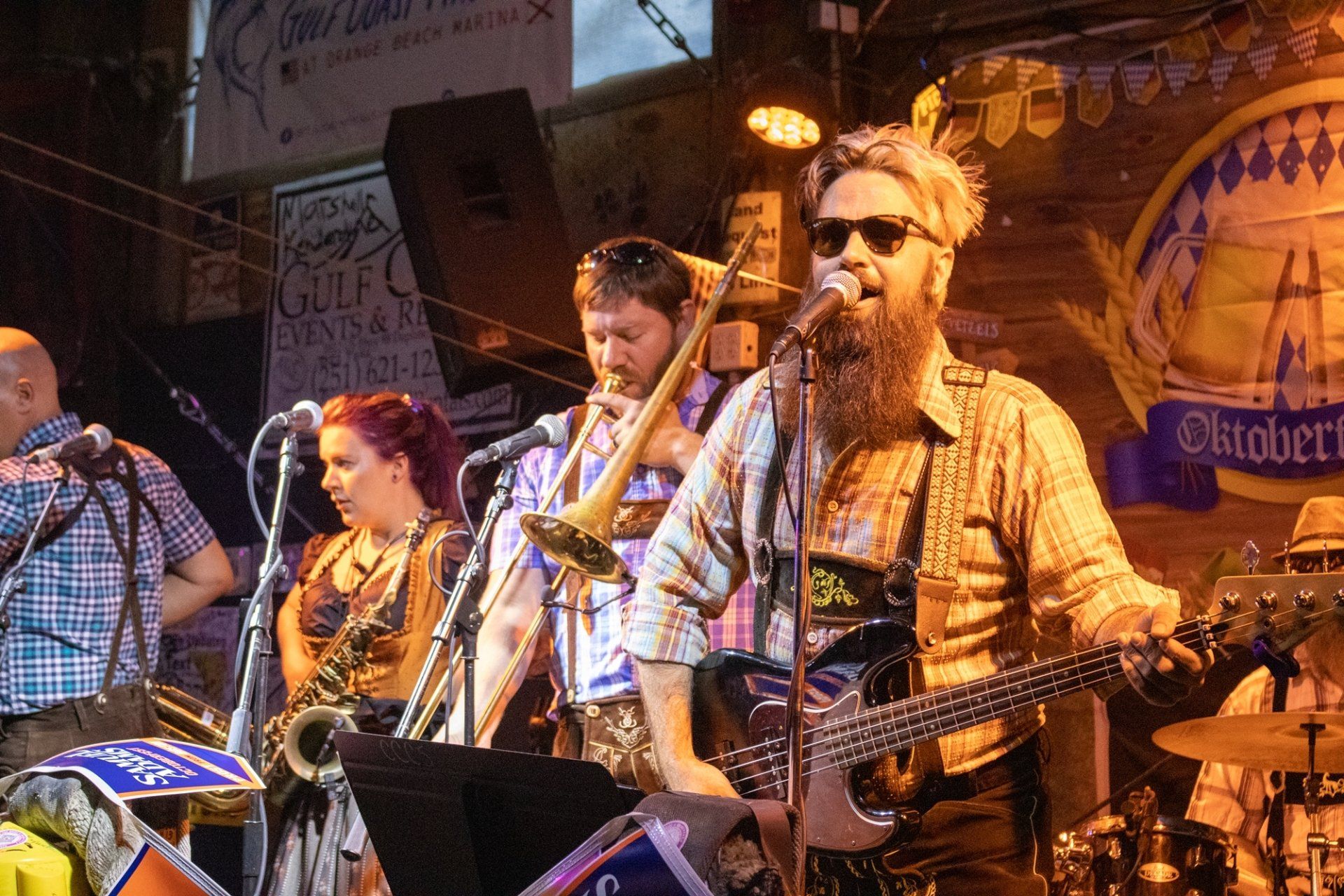 A band wearing lederhosen performs on stage at an Oktoberfest-themed event, featuring a bassist, a horn player, and a saxophonist.