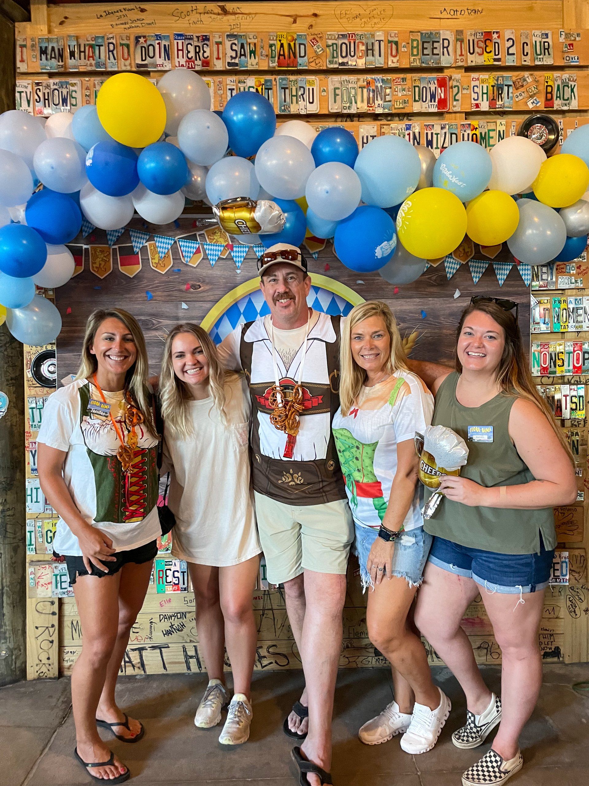 Five people pose for a group photo in front of a wall decorated with license plates and a balloon arch, smiling outdoors.