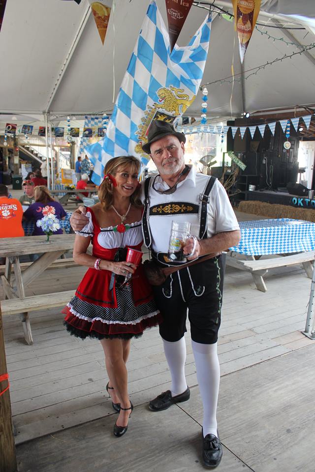 A couple in traditional Bavarian attire poses together under a blue and white checkered flag at an Oktoberfest event.