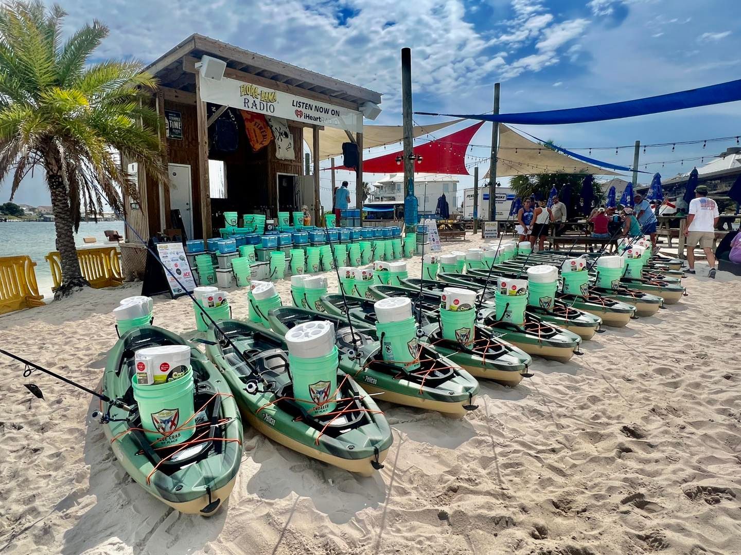 A row of kayaks with light green coolers parked on a sandy beach in front of a waterfront rental shop under a blue sky.