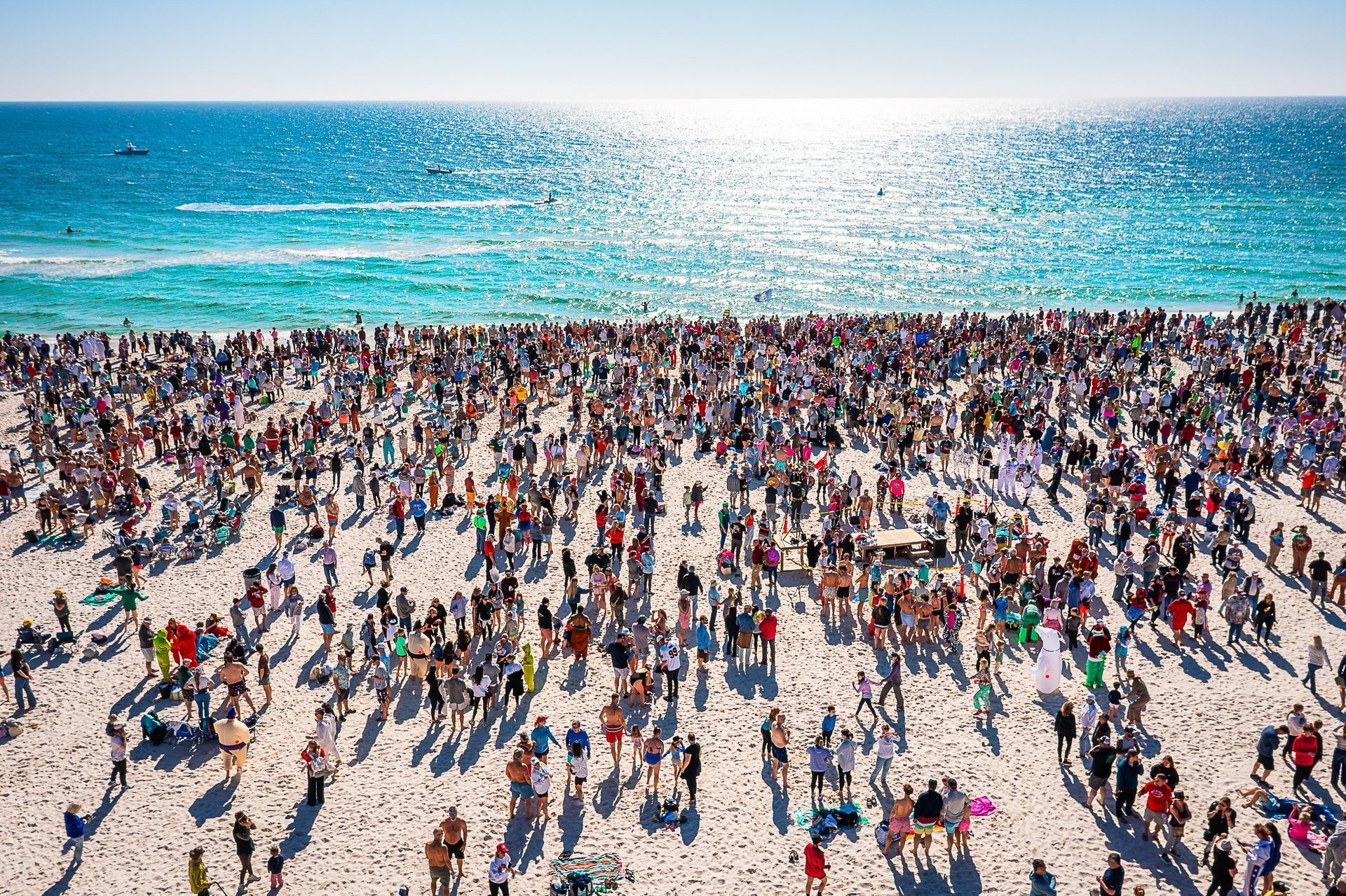 A dense crowd of people filling a bright, sunlit white sand beach bordering a vast, turquoise ocean.