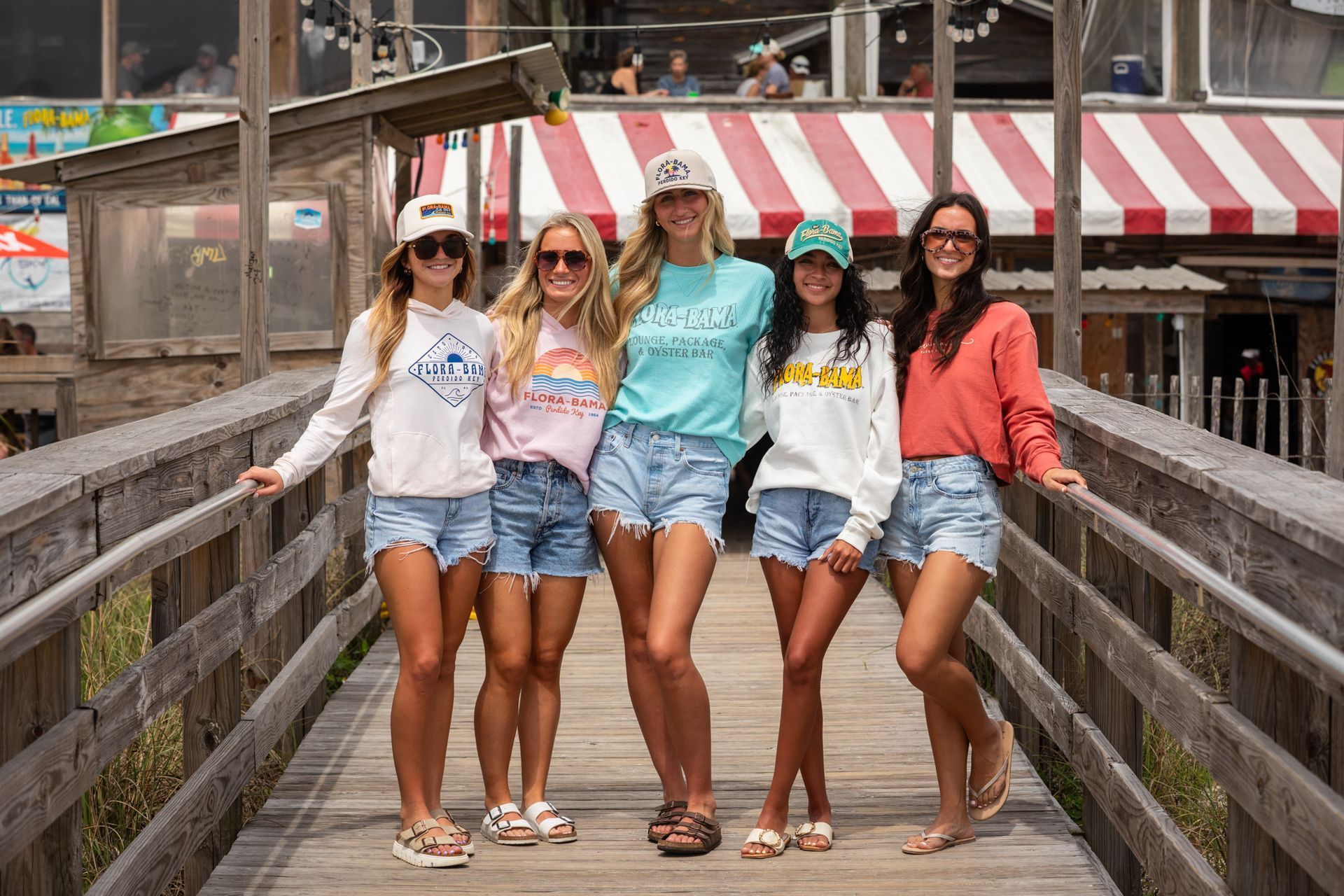 Five people in casual clothing and baseball caps stand together on a wooden pier in front of a building with red awnings.