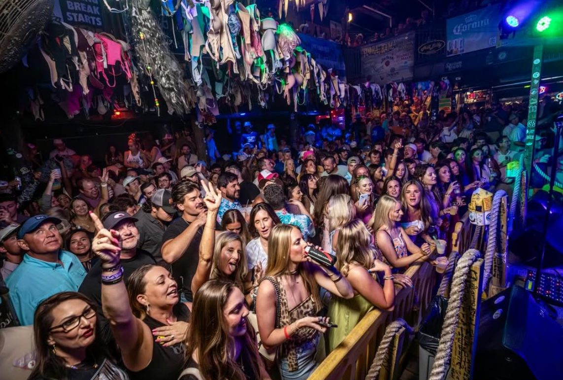 A crowded, dimly lit bar with many people dancing and singing, with dozens of bras hanging from the ceiling rafters.