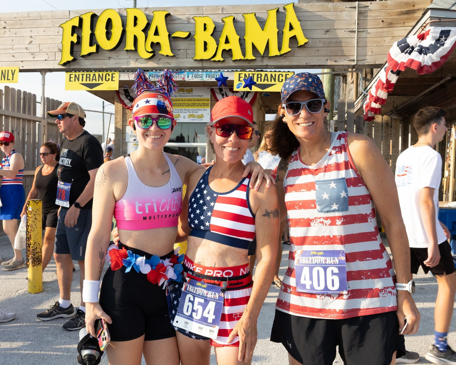 Three women wearing patriotic attire pose in front of a Flora-Bama sign at an outdoor race event.