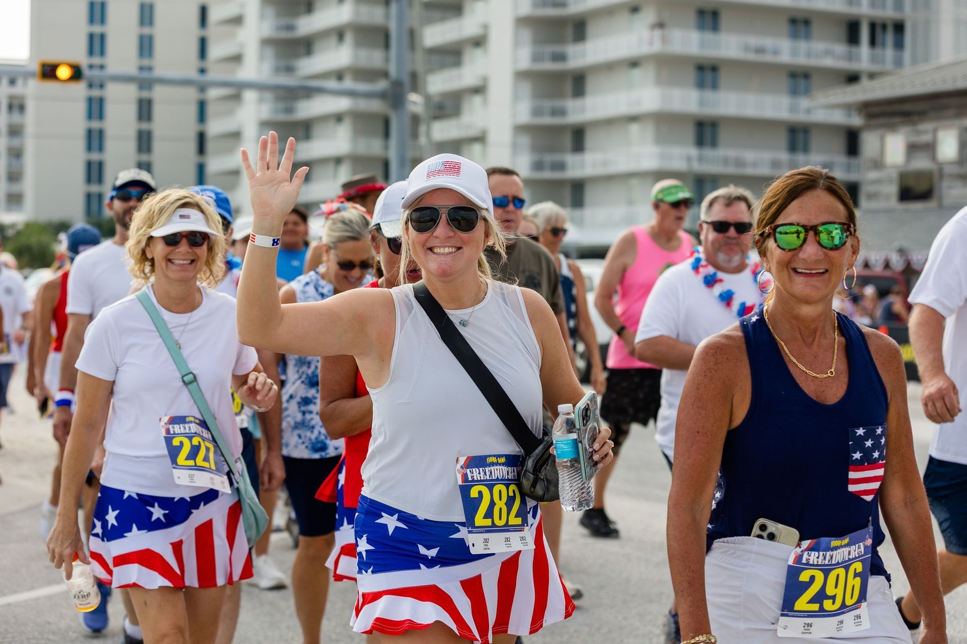Participants in patriotic American-flag-themed running attire wave and walk together during an outdoor race.