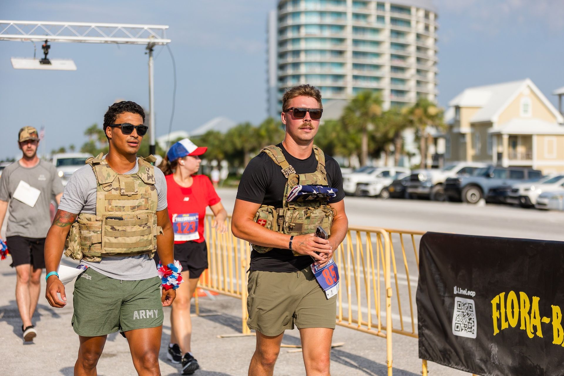 Two participants in weight vests walking past a Flora-Bama banner at an outdoor athletic event.