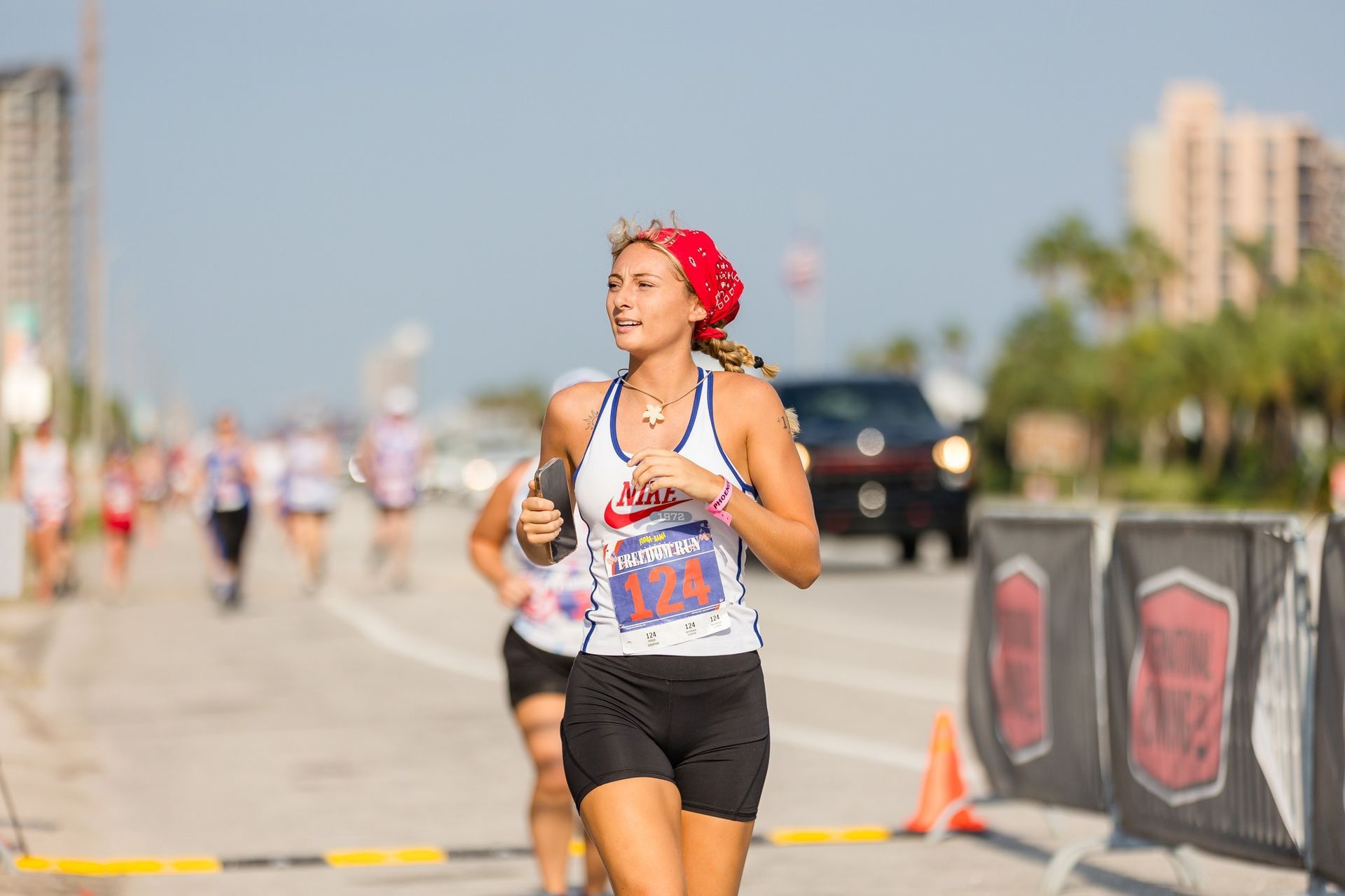 A runner wearing a red bandana and bib number 124 runs on a paved road during a race, with other runners in the background.