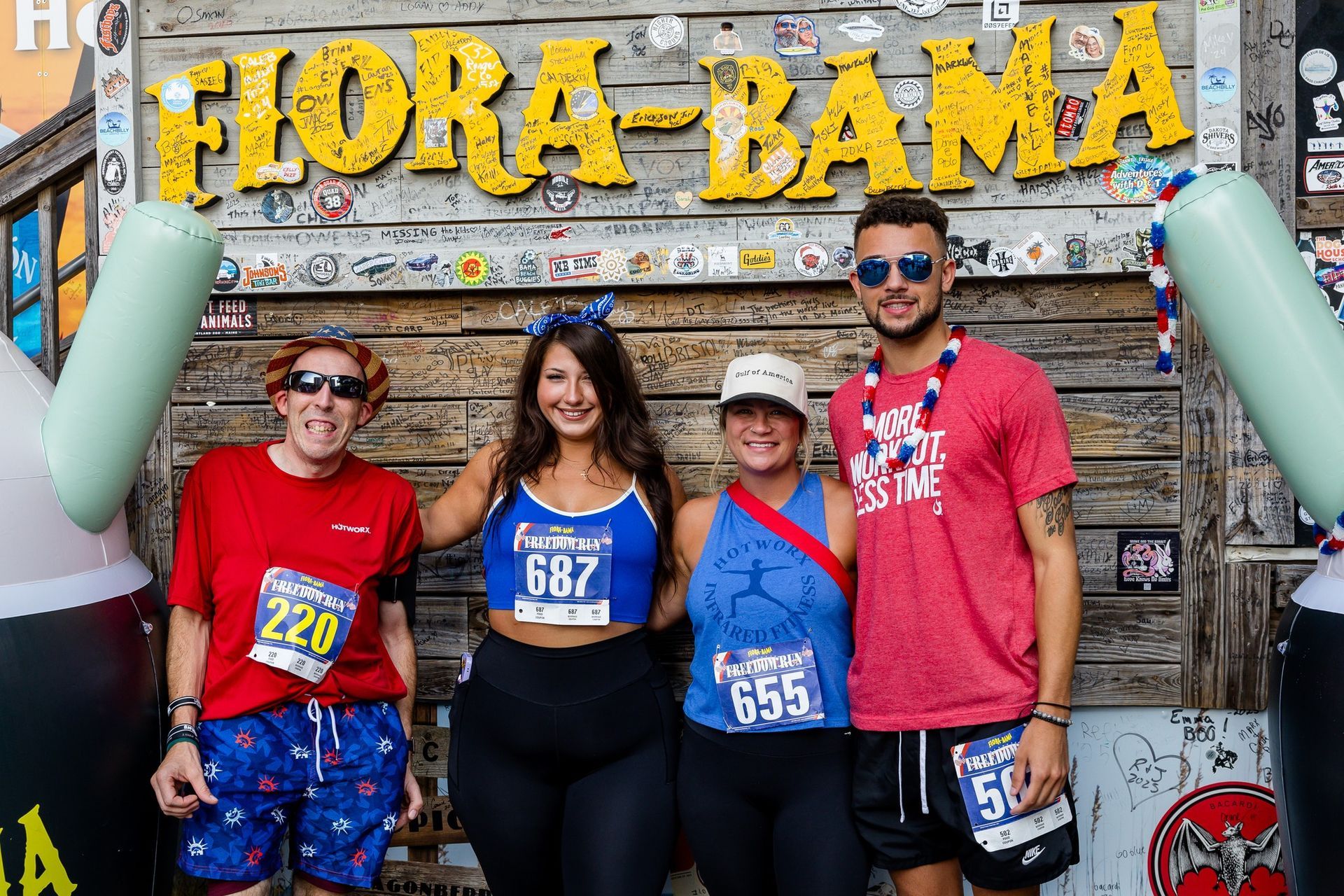 Four people in athletic wear with race bibs pose smiling in front of a wood wall with a large Flora-Bama sign.