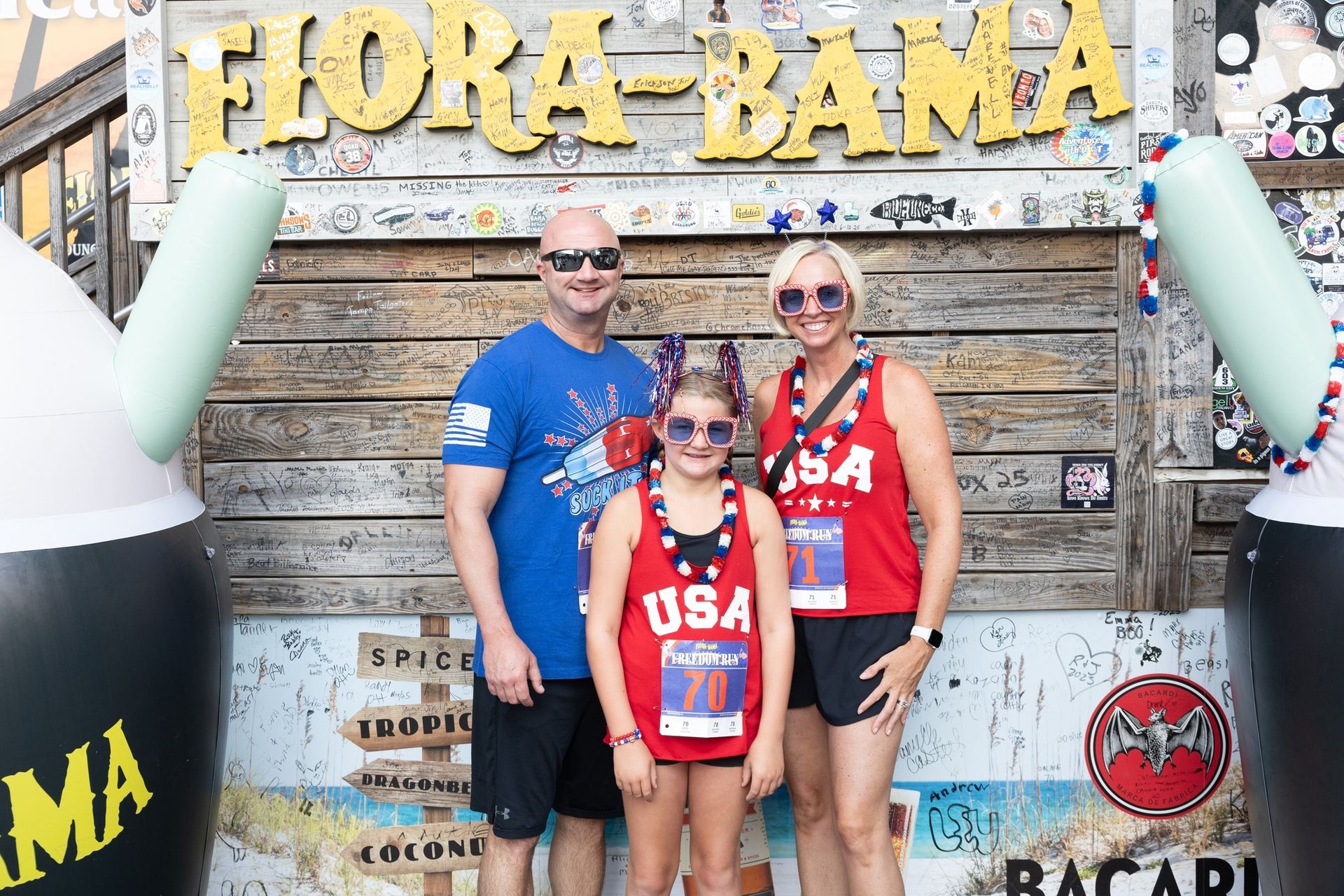 A family wearing patriotic clothing and sunglasses poses in front of a Flora-Bama sign for a festive photograph.
