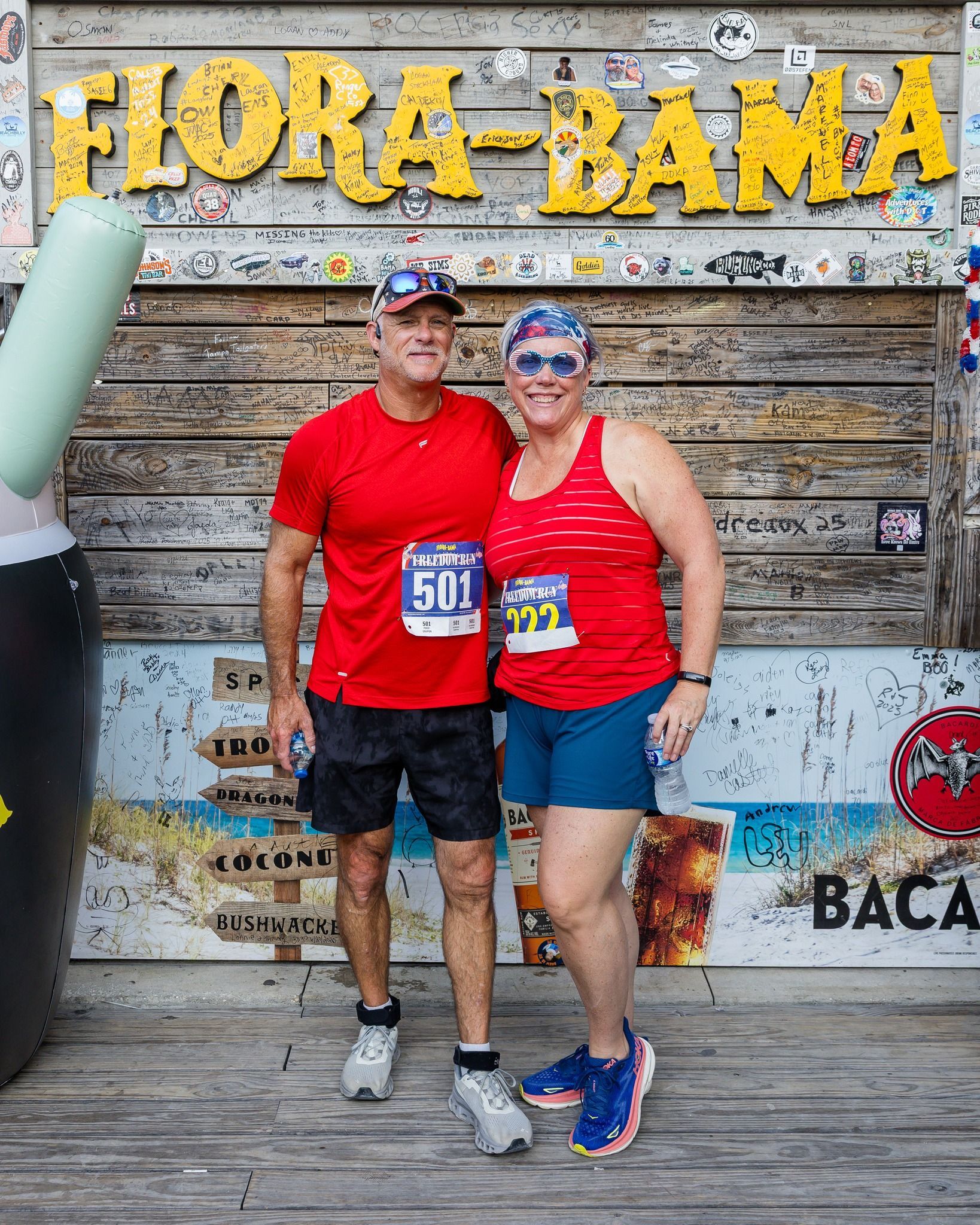 Two runners wearing matching red shirts and bibs smile in front of a Flora-Bama wooden sign.