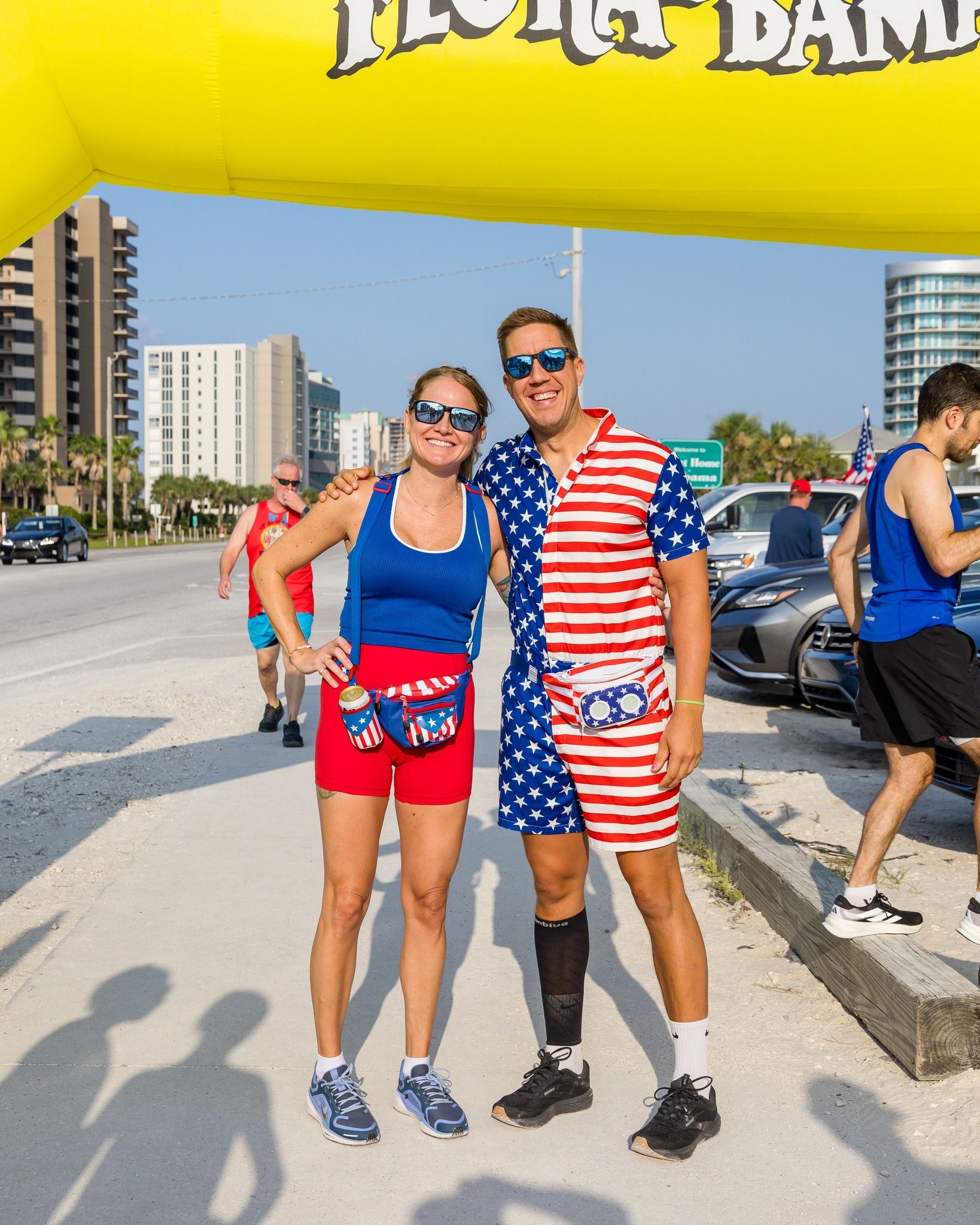 Two people in patriotic-themed outfits stand under a yellow banner on a street, with buildings in the background.