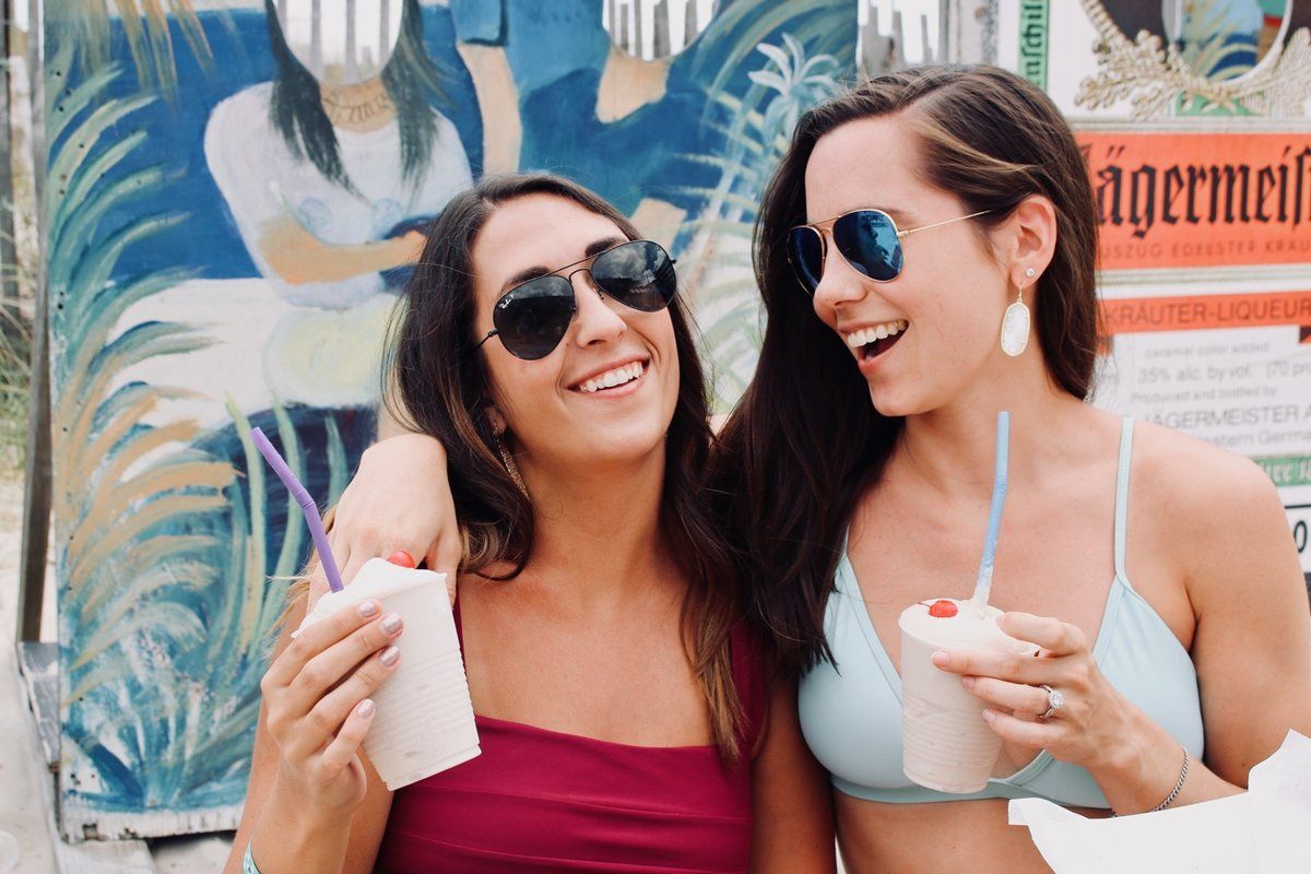 Two friends with sunglasses and drinks smile together in front of a colorful, painted backdrop.