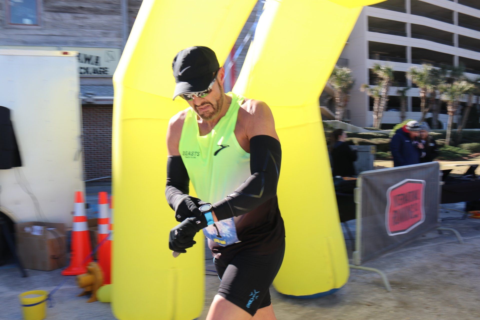 A runner in a neon green tank top and black arm sleeves checks their watch while walking past a yellow inflatable arch.