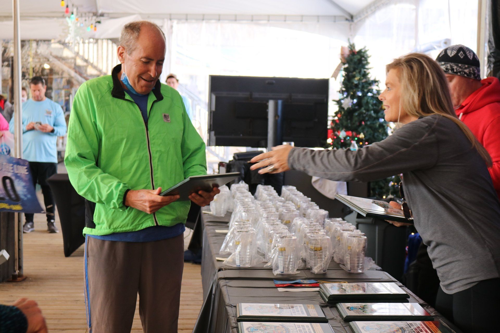 A person in a bright green jacket reviews a digital tablet while gesturing to a woman behind a table of awards.