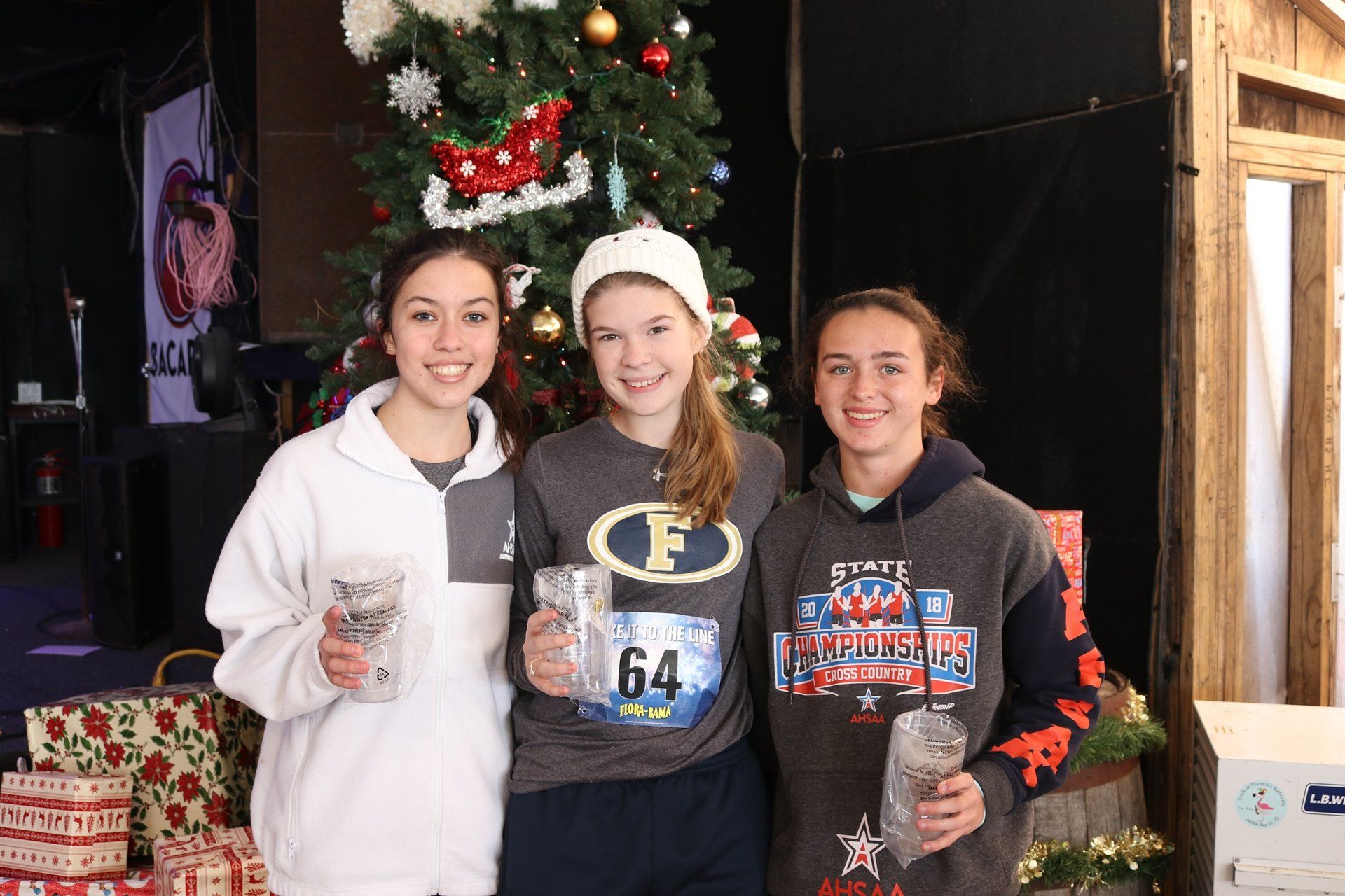 Three people smiling in front of a Christmas tree, holding awards, with one wearing a race bib numbered 64.