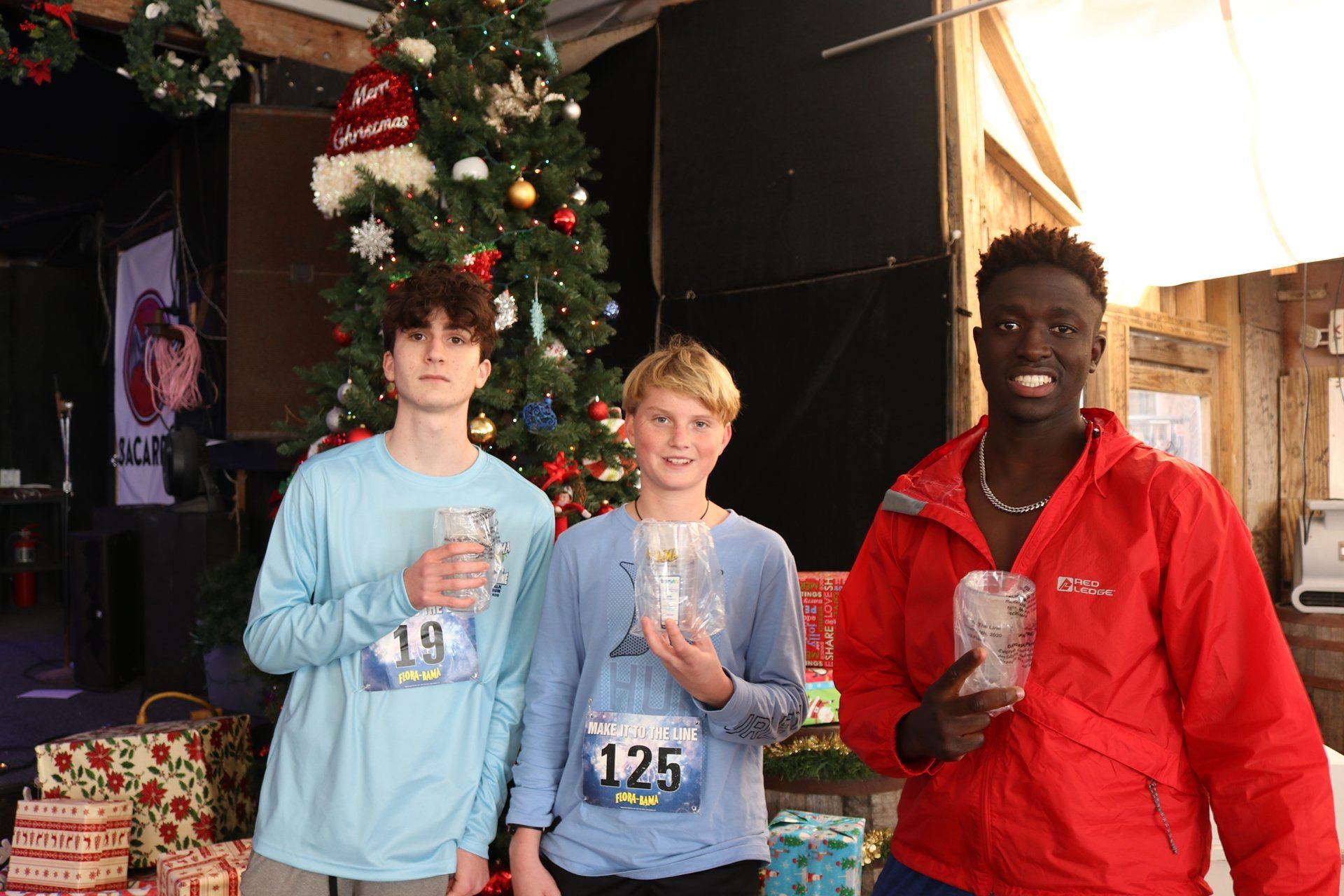 Three participants holding small trophies stand in front of a Christmas tree.