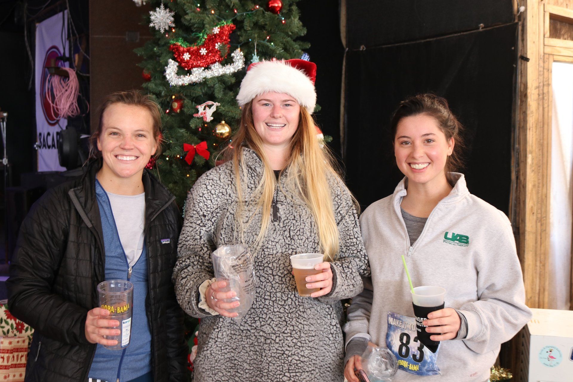 Three people smiling and holding plastic cups in front of a decorated Christmas tree.