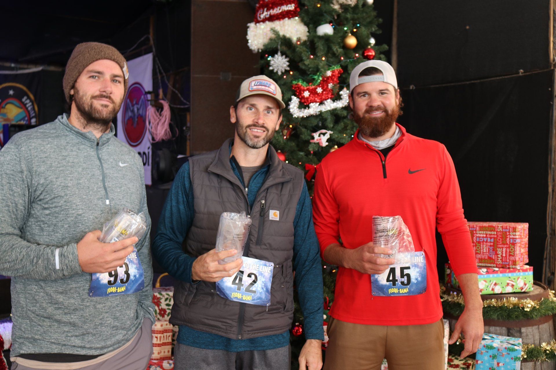 Three individuals with race bibs stand in front of a Christmas tree, each holding a small prize.