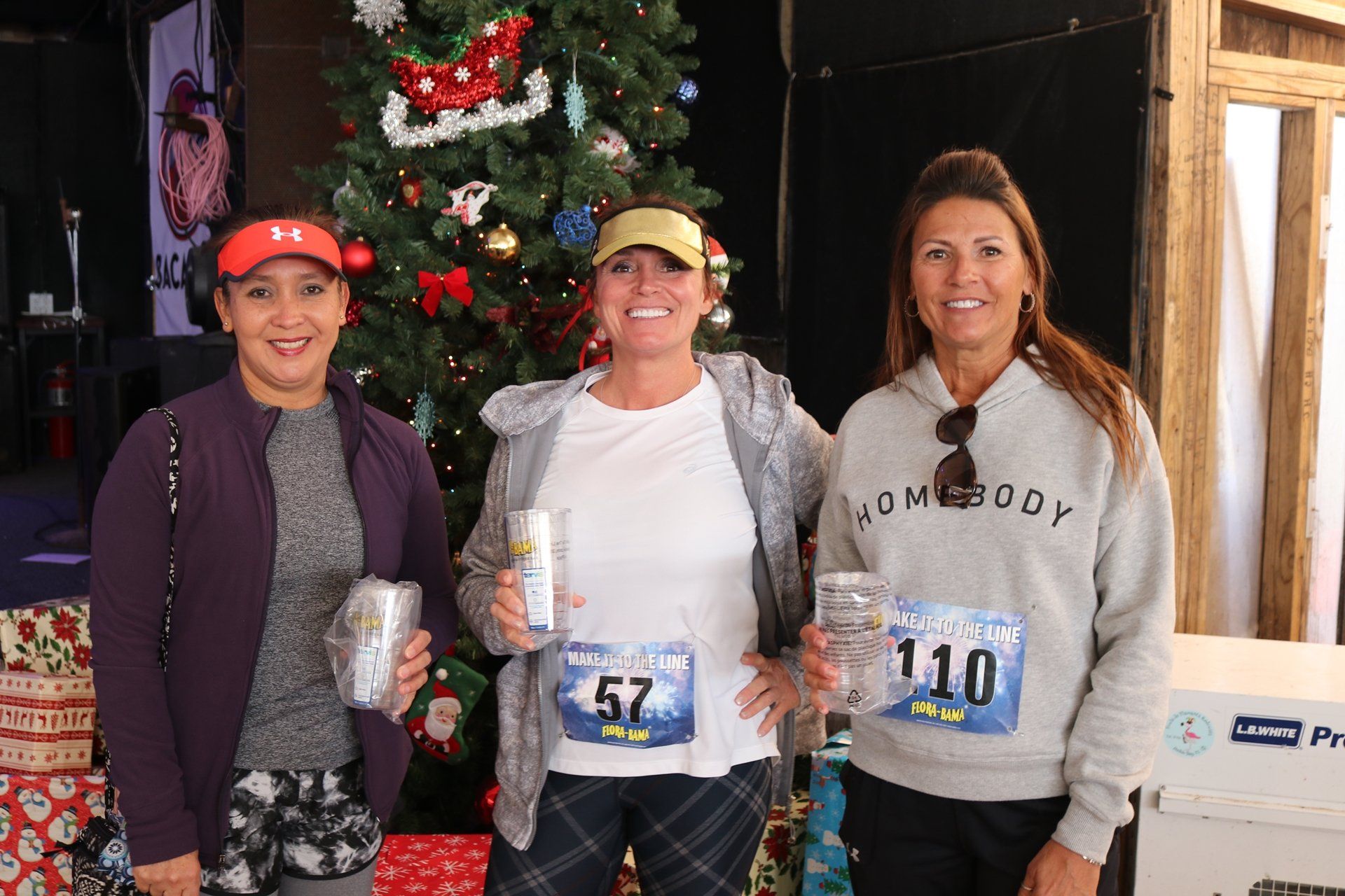 Three women stand in front of a Christmas tree holding awards, wearing race bibs numbered 57 and 110.